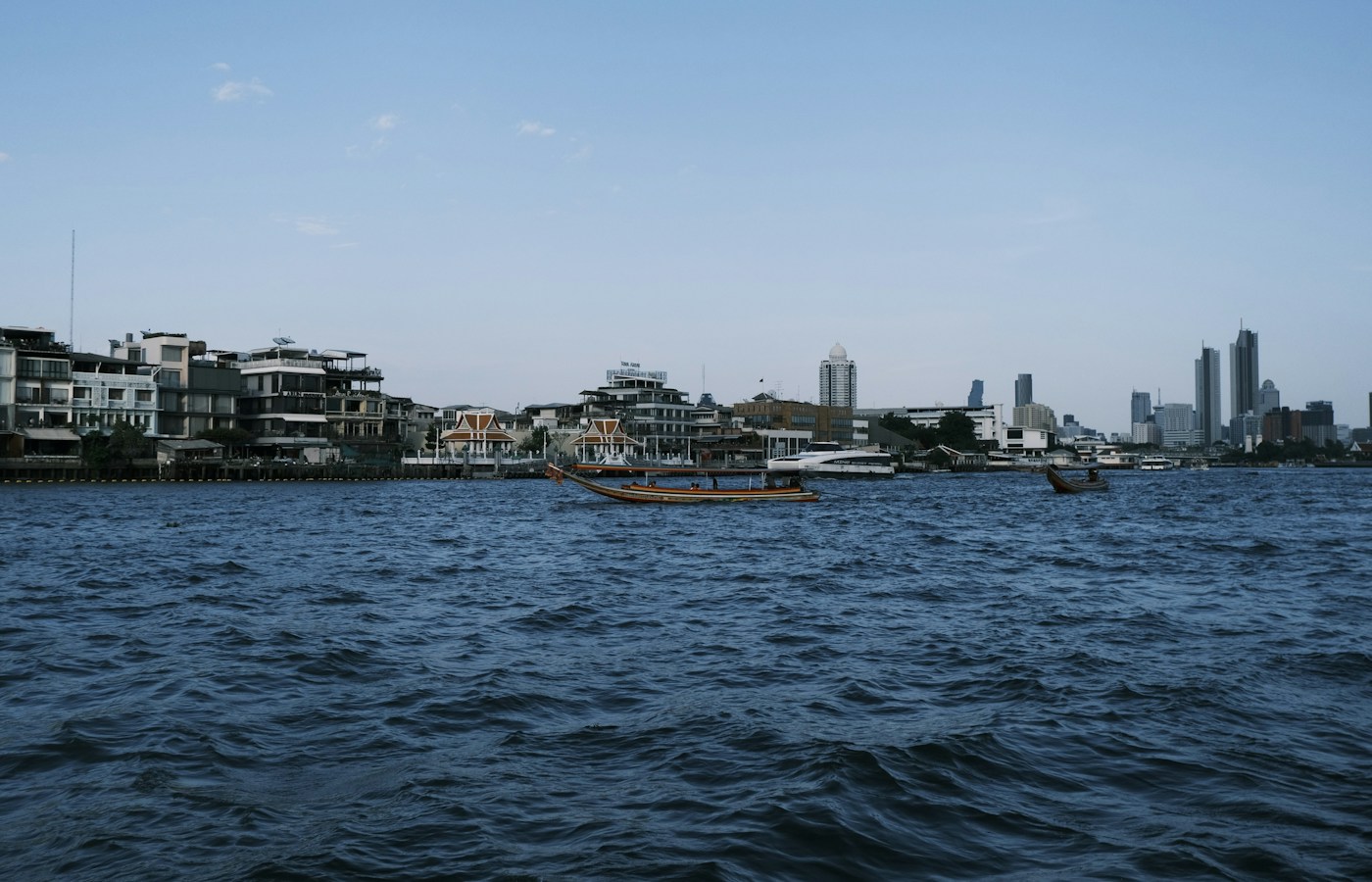 Long-tail boats crossing the Chao Phraya River beneath a blue sky, with Bangkok buildings along the far bank