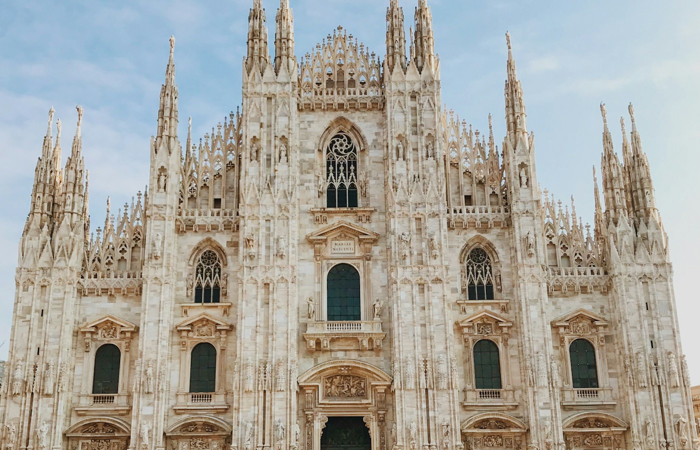 The ornate façade of Milan Cathedral with Gothic spires beneath a partly cloudy sky